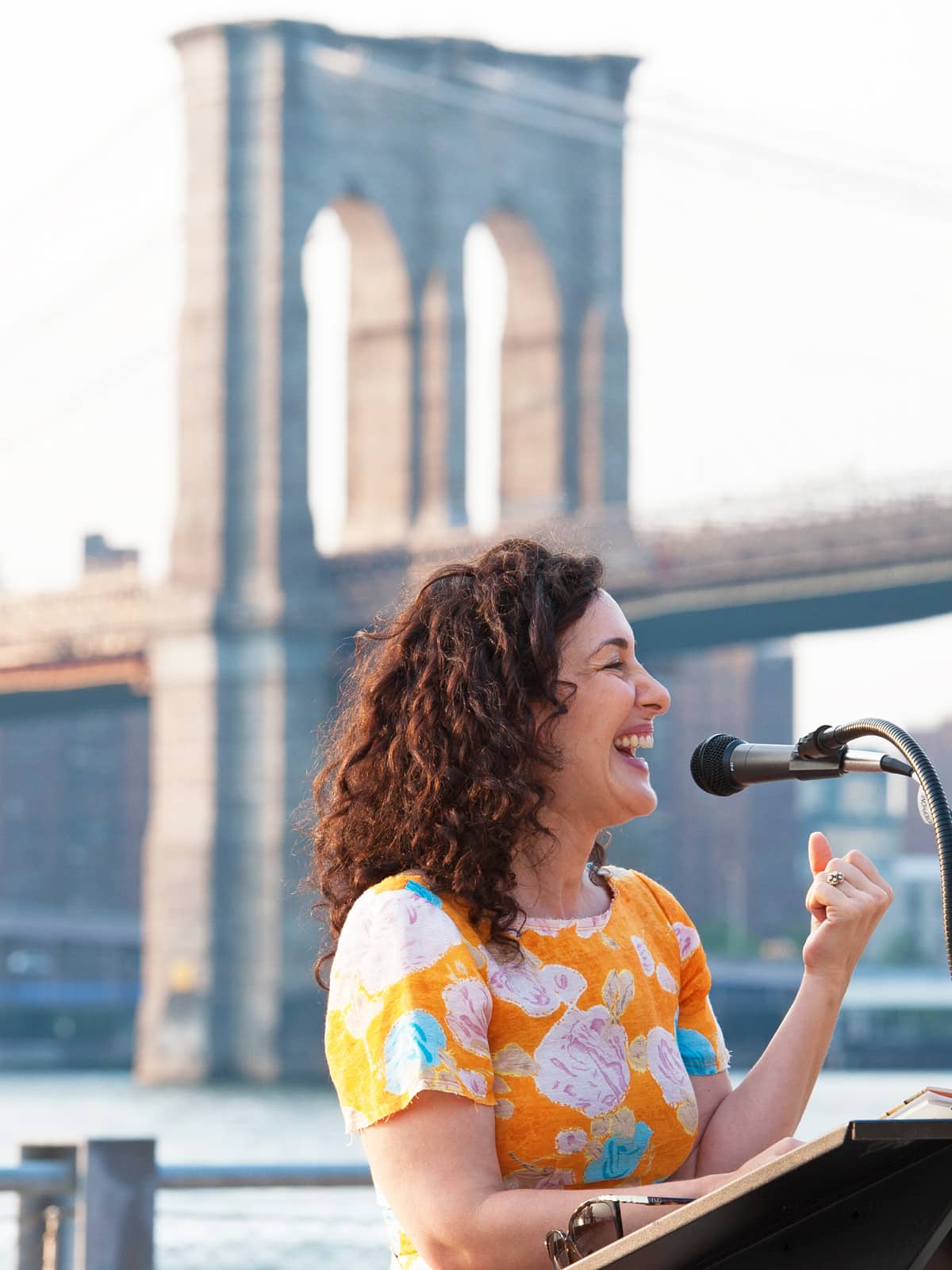 Woman smiling and standing at a podium at sunset. The Brooklyn Bridge is seen in the background.