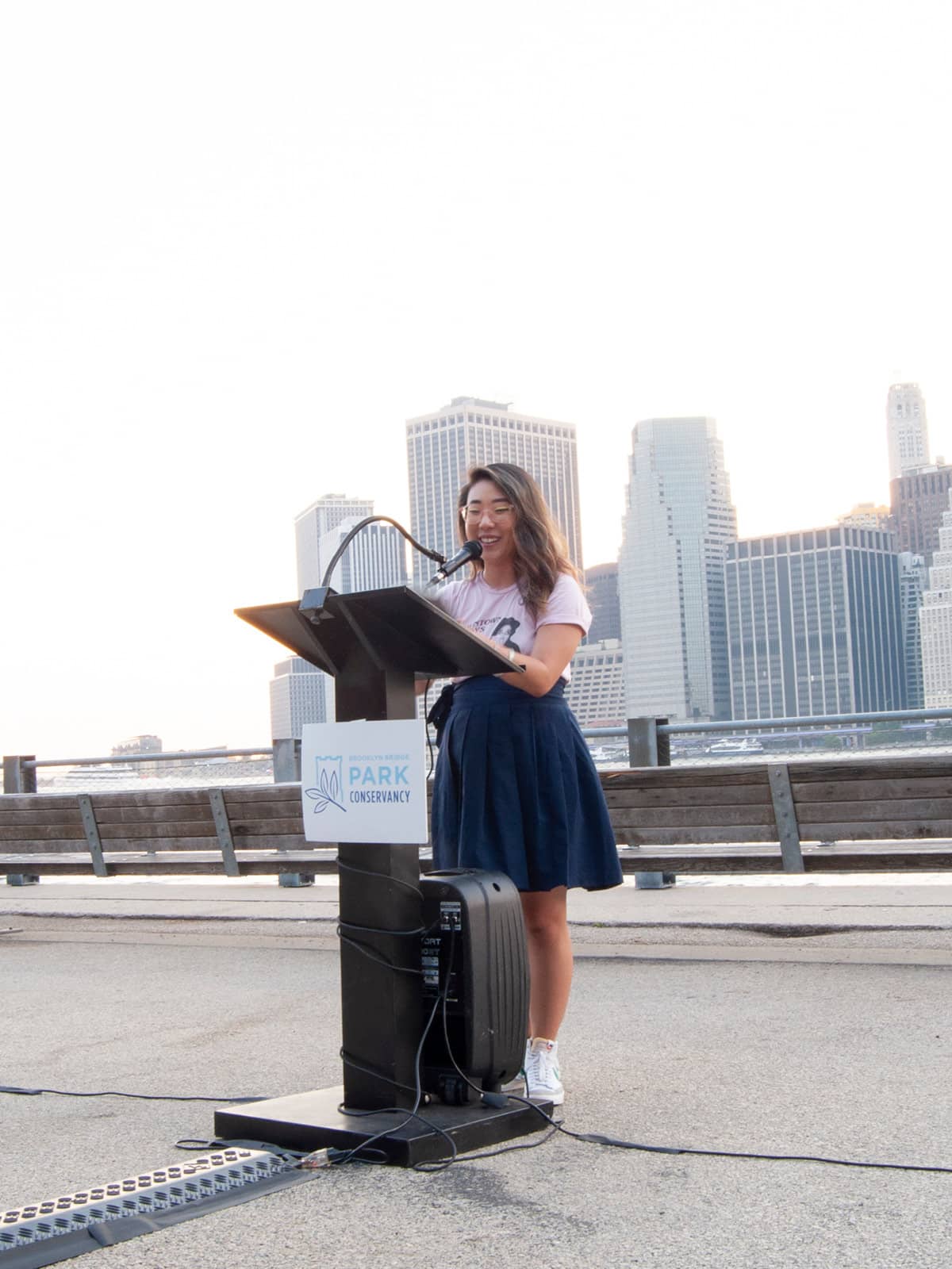 Woman standing at a podium reading out loud at sunset. Lower Manhattan is seen in the background.