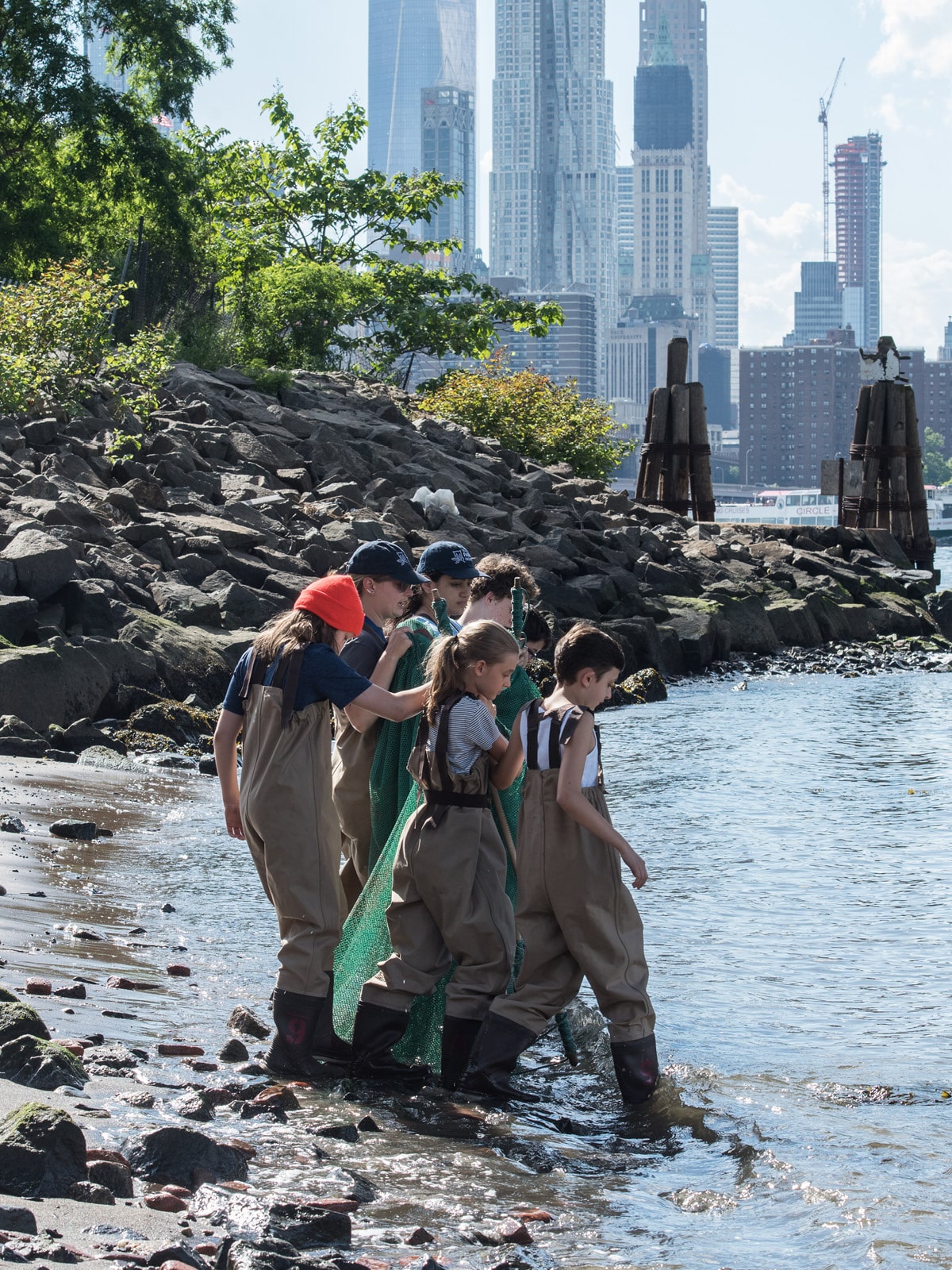 Group of children in fishing waders walking into the water with a large net for seine fishing on a sunny day.