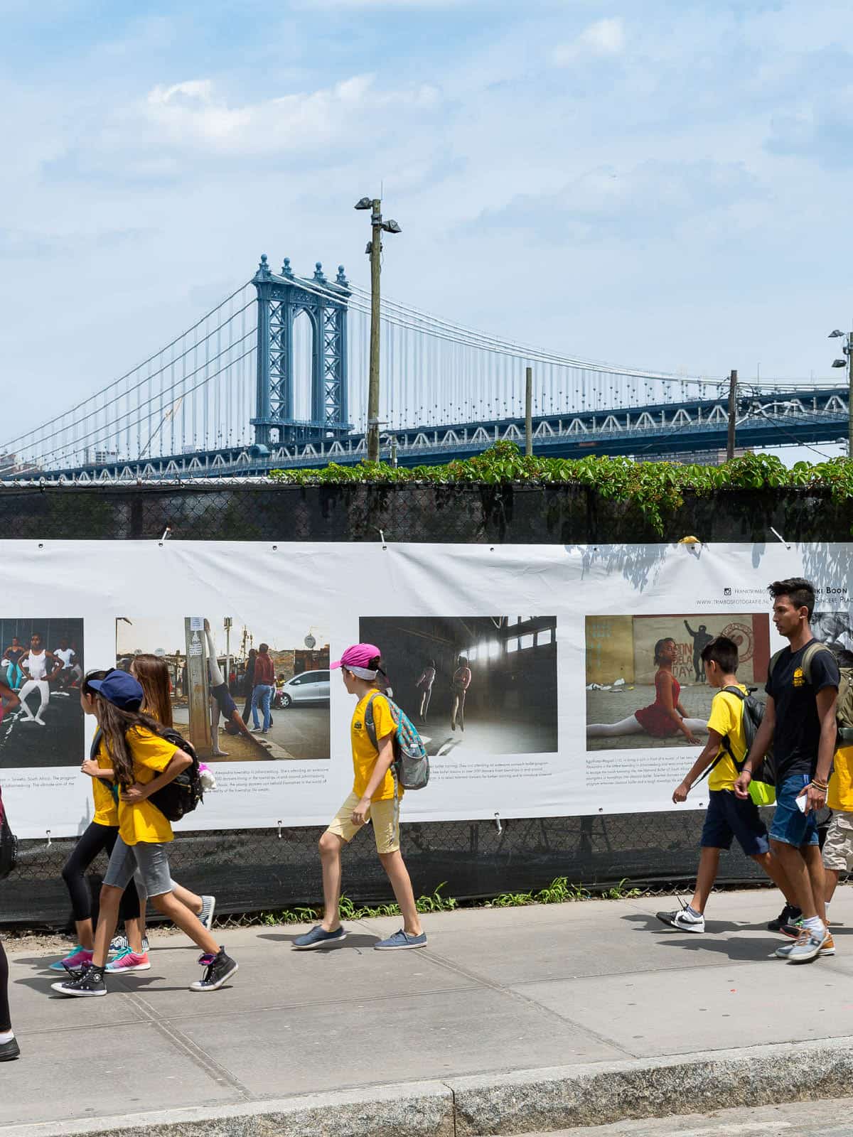 Children walking by a photo banner attached to a chain link fence on a sunny day.