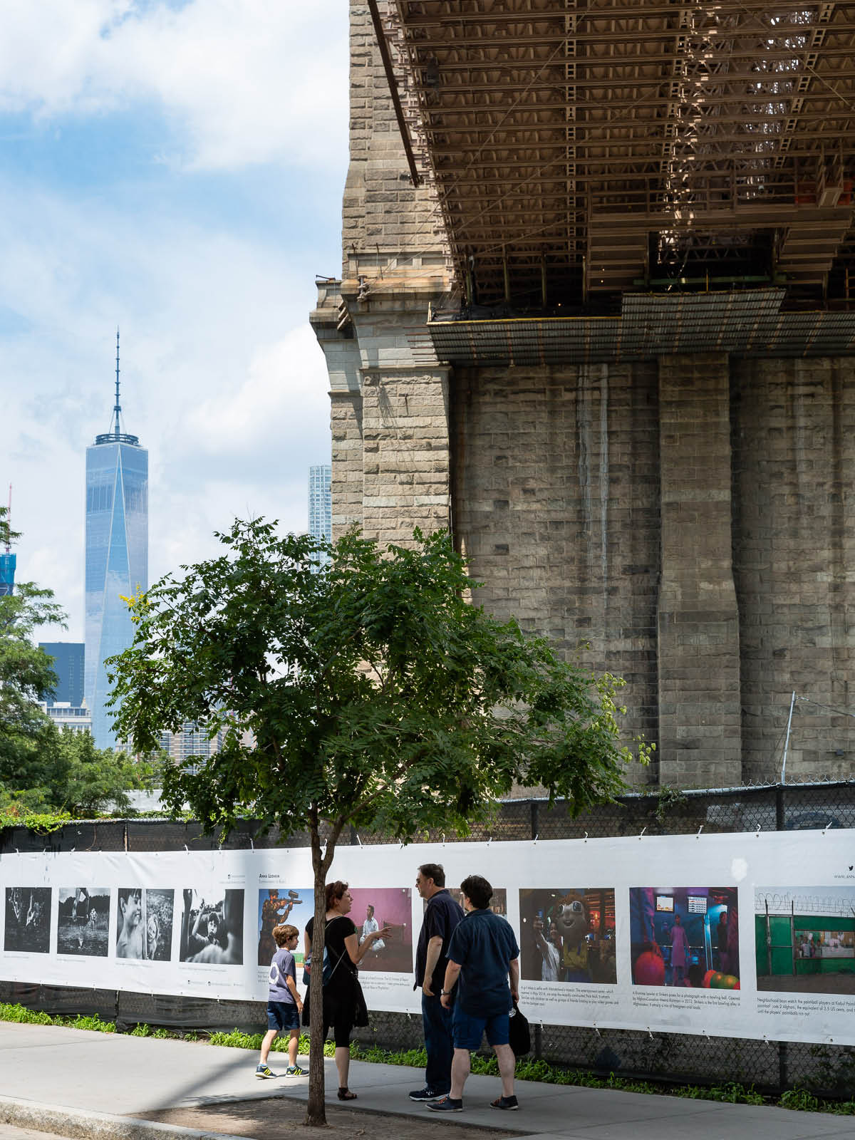 Family looking at photographs on THE FENCE underneath the Brooklyn Bridge.