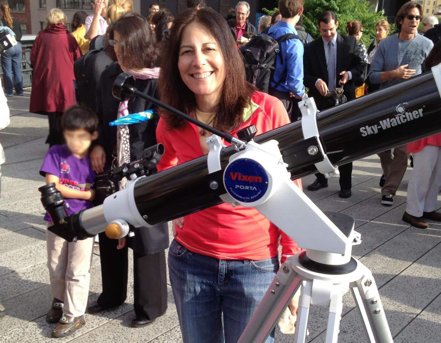 Woman posing with a large telescope on a sunny day.