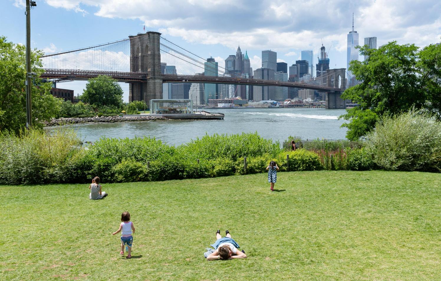People lying on a lawn on a sunny day. The Brooklyn Bridge is seen in the background.