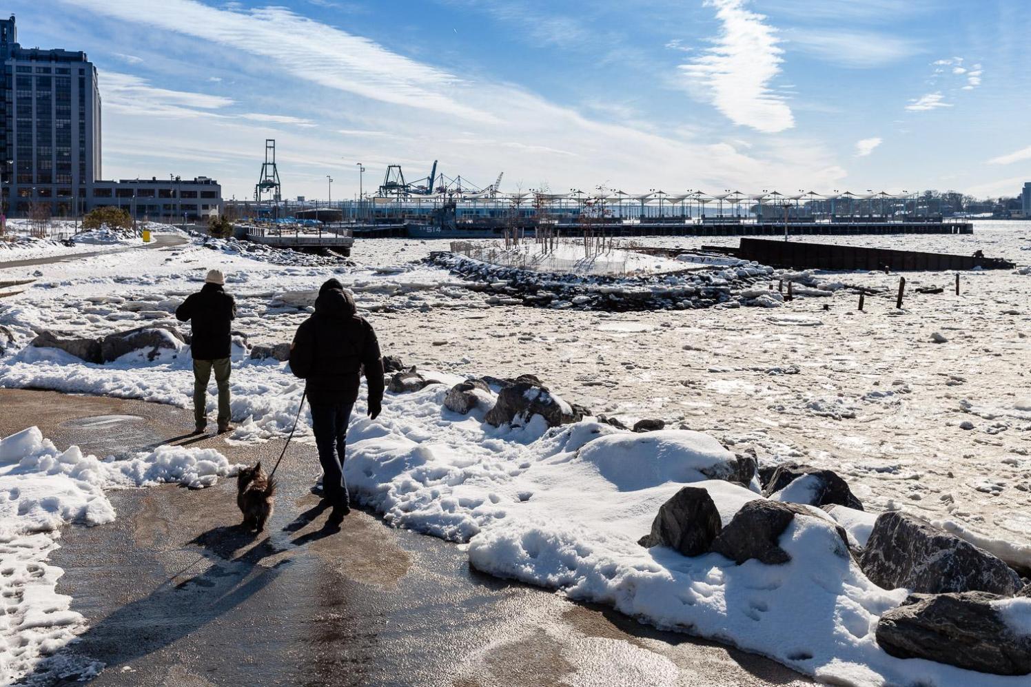 People walking along a path covered in snow by the water on a sunny day.